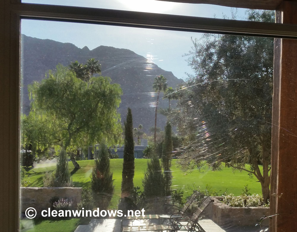 The sun shines on scratches on a glass window with a view of mountains, palm trees, a grass lawn and patio.