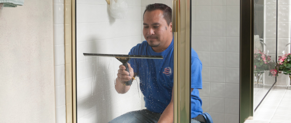 A window cleaner in a blue shirt uses a squeegee on a shower door. 