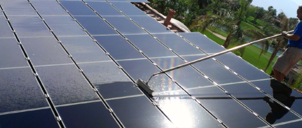 A window cleaner in a bright blue shirt cleans solar panels with a brush on a long stick. 