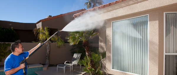 Outside, a window cleaner in a bright blue shirt points a pressure washer high to clean stucco on a home. 